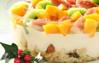 Fotografia em tons de amarelo, verde e vermelho em uma bancada de madeira verde clara, um pano branco com listras verdes, um recipiente de vidro com o pavê de panetone com frutas dentro dele. Fotografia em tons de amarelo, verde e vermelho em uma bancada de madeira verde clara, um pano branco com listras verdes, um recipiente de vidro com o pavê de panetone com frutas dentro dele.
