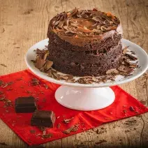 Foto da receita de Bolo Brownie com Brigadeiro. O bolo em tom marrom escuro está em cima de um prato branco mais alto, sobre uma bancada de madeira decorada com raspas de chocolate