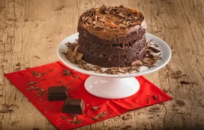 Foto da receita de Bolo Brownie com Brigadeiro. O bolo em tom marrom escuro está em cima de um prato branco mais alto, sobre uma bancada de madeira decorada com raspas de chocolate