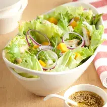 em uma mesa de madeira contém um recipiente branco que comporta a salada. Ao lado um pote com tempero e um pano listrado nas cores branco e rosa.