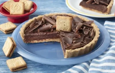 Fotografia em tons de azul em uma bancada de madeira azul, um pano azul listrado, um prato redondo raso azul com a torta de Biscoito Passatempo em cima dele, já com uma fatia cortada. Ao fundo, um prato com a fatia da torta e um potinho com os biscoitos.