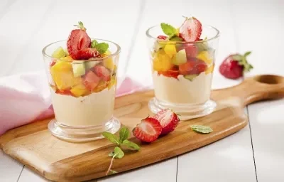 Fotografia em tons de branco, marrom e rosa de uma bancada de madeira branca com uma tabua de madeira e sobre ela dois o copos de vidro redondos com a salada de frutas especial e fatias de morangos espalhadas. Ao fundo um paninho rosa claro. Fotografia em tons de branco, marrom e rosa de uma bancada de madeira branca com uma tabua de madeira e sobre ela dois o copos de vidro redondos com a salada de frutas especial e fatias de morangos espalhadas. Ao fundo um paninho rosa claro.