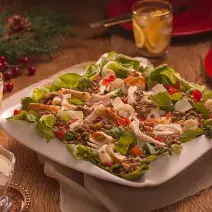 Fotografia em tons de vermelho em uma bancada de madeira escura, um pano bege, um prato branco grande com salada de alface, rúcula, lentilha e chester. Ao lado, copos, molho e enfeites e decoração natalina.