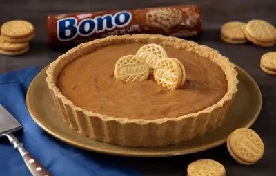 Foto de uma torta de doce de leite de cores marrom clara e escura. Ela está sobre um prato marrom, numa mesa com um tecido azul e uma espátula de bolo de aço inox. Há biscoitos Bono Doce de Leite espalhados e ao fundo um pacote de Biscoito Bono Foto de uma torta de doce de leite de cores marrom clara e escura. Ela está sobre um prato marrom, numa mesa com um tecido azul e uma espátula de bolo de aço inox. Há biscoitos Bono Doce de Leite espalhados e ao fundo um pacote de Biscoito Bono