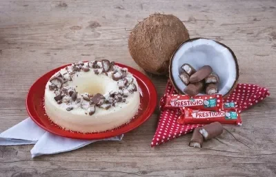 Foto em tons de vermelho e marrom da receita de pudim de prestígio servida em um prato de porcelana vermelho com panos brancos embaixo. Ao lado, um coco cortado ao meio com prestígios dentro e mais embalagens a frente, além de um pano vermelho com bolas