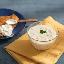 Fotografia em tons de azul, bege e branco com bancada azul contendo prato azul com porção de peixe empanado e molho com uma colher, ao lado potinho com um molho branco sobre guardanapo bege.
