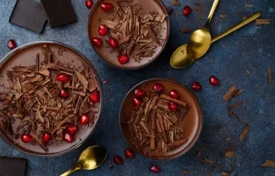 Fotografía oscura tomada desde arriba de tres grandes recipientes de vidrio sobre una mesa oscura, que contienen cucharas doradas, virutas y trozos de chocolate. Dentro de los recipientes se encuentra el postre, cubierto con ganache y virutas.