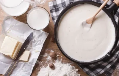 Fotografia em tons de marrom com uma panela preta ao centro. Dentro da panela existe um molho branco. Ao lado existe uma manteiga em tablete cortada em fatias. Fotografia em tons de marrom com uma panela preta ao centro. Dentro da panela existe um molho branco. Ao lado existe uma manteiga em tablete cortada em fatias.