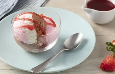 Imagen en primer plano de la receta de helado de moça Napolitano servida en un recipiente de vidrio, decorada con un almíbar rosa, todo en un plato azul sobre un mostrador claro con fresas enteras y cubiertos. Imagen en primer plano de la receta de helado de moça Napolitano servida en un recipiente de vidrio, decorada con un almíbar rosa, todo en un plato azul sobre un mostrador claro con fresas enteras y cubiertos.