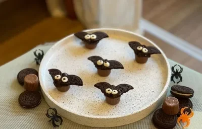 Fotografía en tonos marrones sobre una encimera de madera blanca. En el centro, un plato redondo blanco con los murciélagos. Cerca hay chocolates y galletas.