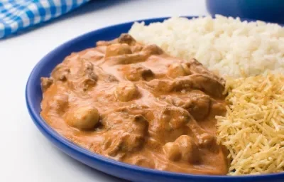 Fotografia em tons de azul com um prato azul ao centro. Dentro do prato existe uma receita com carne, champignons e molho rose. Ao lado existe uma porção e batata palha. Fotografia em tons de azul com um prato azul ao centro. Dentro do prato existe uma receita com carne, champignons e molho rose. Ao lado existe uma porção e batata palha.