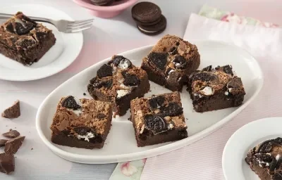 Foto de la receta de Brownie con Leche Condensada y Galletas Negresco. Muestra 7 rebanadas de brownie cortadas y dispuestas en un molde ovalado blanco sobre un fondo rosa pastel.