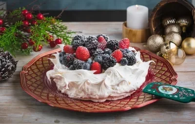 Foto de una receta de pavlova navideña blanca, en un plato rojo, decorada con frutos rojos y azúcar. Está sobre una mesa de madera llena de adornos navideños como piñas, bolas de Navidad y una vela encendida al fondo.