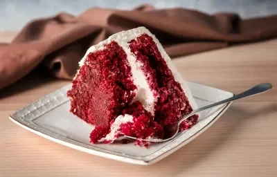 Fotografía de una porción de pastel red velvet sobre un plato de cerámica blanca, con glaseado de queso crema y un tenedor plateado al lado. El fondo es una tela marrón suave sobre una mesa de madera clara.