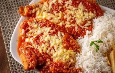 Fotografía de un plato rectangular blanco con dos filetes de pollo a la parmesana cubiertos con queso fundido, acompañados de arroz y patatas fritas.