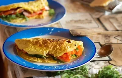 Fotografía en tonos azules y naranjas sobre una encimera de madera oscura. Un plato redondo azul con una tortilla rellena de tomate y rúcula encima. Junto a él, una cuchara y unas servilletas marrones estampadas.