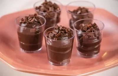 Fotografia em tons de rosa com 5 copinhos de plástico ao centro. Dentro de capa copo existe um brigadeiro de colher de cor marrom escura.