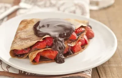 Fotografía en tonos marrones sobre una encimera de madera con un mantel blanco con rayas marrones, un plato blanco poco profundo con la crepe rellena de fresas y chocolate. Fotografía en tonos marrones sobre una encimera de madera con un mantel blanco con rayas marrones, un plato blanco poco profundo con la crepe rellena de fresas y chocolate.