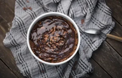 Fotografía de un pastel de avena horneado y frito al aire en un ramequín blanco sobre un paño gris arrugado, colocado sobre una tabla de madera oscura. Se vierte jarabe de chocolate sobre el pastel.