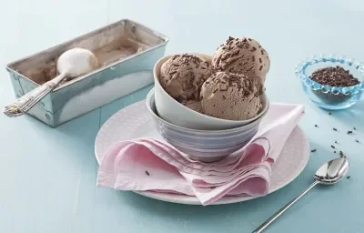 Fotografía en tonos azules y rosas sobre un banco de madera celeste, un paño rosa, un pequeño plato redondo rosa con potes de porcelana encima y bolitas de helado de brigadeiro con chispitas para decorar. Al lado, un botecito con chispitas y una cuchara.