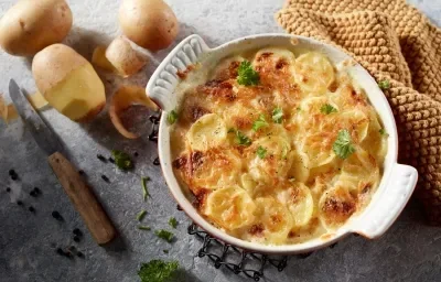 Foto da receita de Batata Gratinada Zero lactose. Observa-se um refratário redondo branco de porcelana com as batatas gratinadas dentro. Ao lado esquerdo, batatas descascadas decoram Foto da receita de Batata Gratinada Zero lactose. Observa-se um refratário redondo branco de porcelana com as batatas gratinadas dentro. Ao lado esquerdo, batatas descascadas decoram