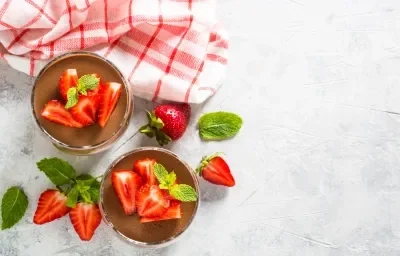 Fotografía en tonos claros, vista superior, de dos vasos decorativos de tamaño estándar que contienen un postre con sabor a fresa, cubierto de chocolate y decorado con trozos de fresas y hojas de menta.