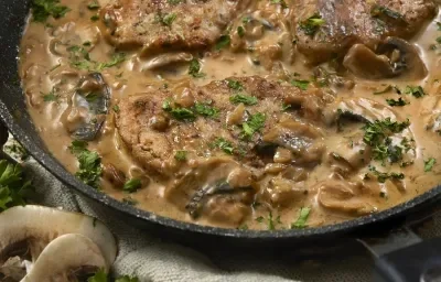 Fotografía en tonos marrones sobre una encimera de madera marrón. En el centro, una sartén negra con los medallones, y junto a ella, algunos palmitos dispersos.