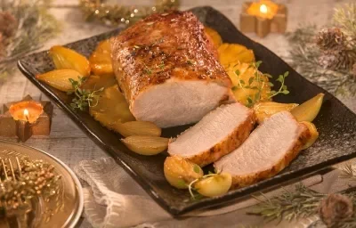Fotografia em tons de amarelo em uma bancada de madeira clara, um recipiente retangular preto com o lombo com os abacaxis caramelizados em volta. Ao lado e atrás, decoração e enfeites natalinos.