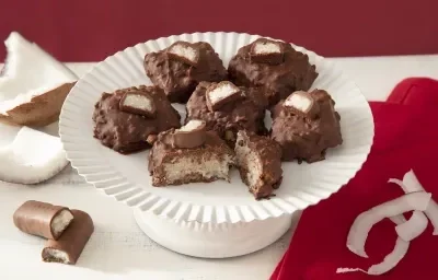 Foto da receita de cocada de prestígio. Observa-se uma boleira branca com 6 cocadas banhadas no chocolate. A da frente está cortada e é possível ver o coco de dentro Foto da receita de cocada de prestígio. Observa-se uma boleira branca com 6 cocadas banhadas no chocolate. A da frente está cortada e é possível ver o coco de dentro