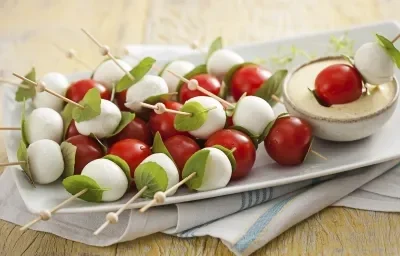 Fotografía en tonos blancos y rojos, en el centro una fuente con brochetas de tomates cherry, mozzarella de búfala y albahaca, junto a una pequeña olla con salsa cremosa, sobre una servilleta blanca con rayas azules en una encimera de madera.