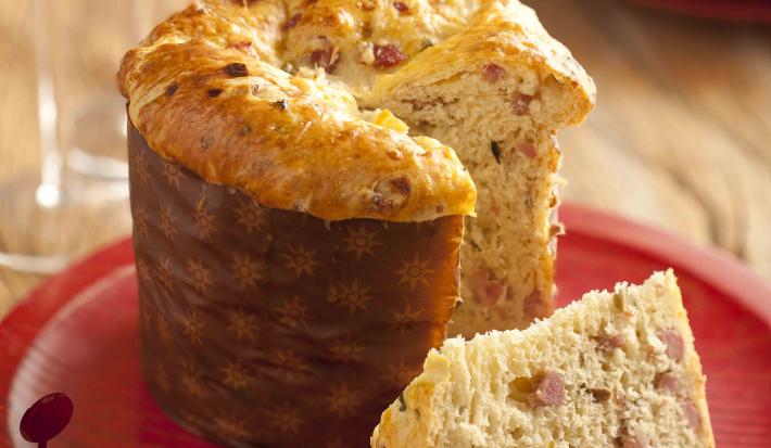 Fotografía en tonos rojos y marrones, en el centro un plato rojo con un panettone en rodajas, al lado de dos vasos con bebidas y una decoración navideña, al fondo dos platos apilados, todo sobre una rústica encimera de madera.