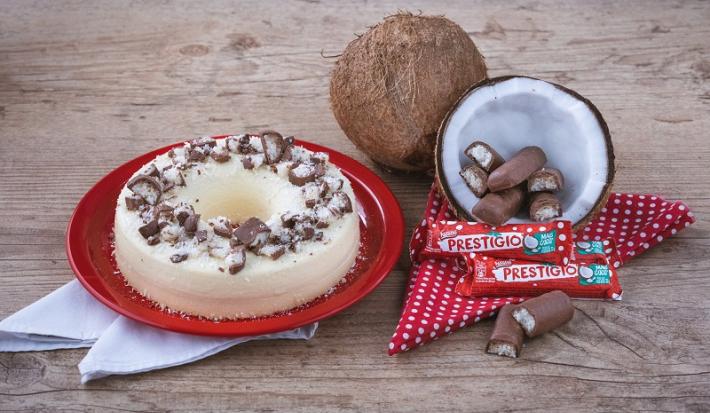 Foto em tons de vermelho e marrom da receita de pudim de prestígio servida em um prato de porcelana vermelho com panos brancos embaixo. Ao lado, um coco cortado ao meio com prestígios dentro e mais embalagens a frente, além de um pano vermelho com bolas