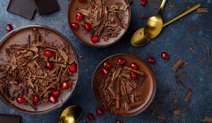 Fotografía oscura tomada desde arriba de tres grandes recipientes de vidrio sobre una mesa oscura, que contienen cucharas doradas, virutas y trozos de chocolate. Dentro de los recipientes se encuentra el postre, cubierto con ganache y virutas.