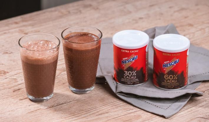 Foto da receita de smoothie pós treino de nescau extra cacau servida em dois copos de vidro altos sobre uma mesa de madeira com duas embalagens de nescau extra cacau 30% e 60% sobre um paninho cinza