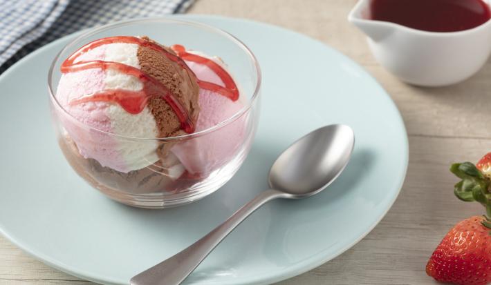 Imagen en primer plano de la receta de helado de moça Napolitano servida en un recipiente de vidrio, decorada con un almíbar rosa, todo en un plato azul sobre un mostrador claro con fresas enteras y cubiertos.