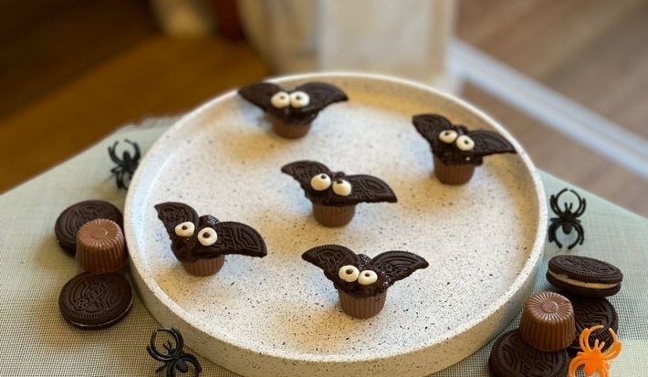 Fotografía en tonos marrones sobre una encimera de madera blanca. En el centro, un plato redondo blanco con los murciélagos. Cerca hay chocolates y galletas.