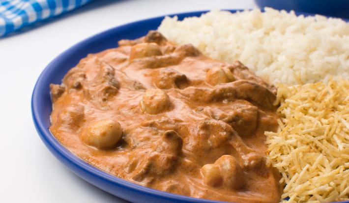 Fotografia em tons de azul com um prato azul ao centro. Dentro do prato existe uma receita com carne, champignons e molho rose. Ao lado existe uma porção e batata palha.