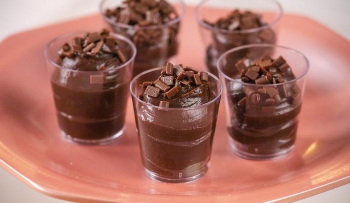 Fotografia em tons de rosa com 5 copinhos de plástico ao centro. Dentro de capa copo existe um brigadeiro de colher de cor marrom escura.