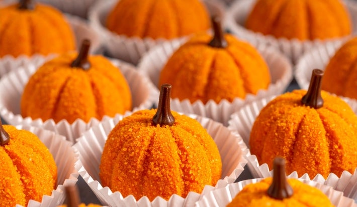 Fotografía en tonos naranjas con varios brigadeiros con forma de calabaza en el centro. Los brigadeiros se elaboran con calabaza cocida, rebozada en azúcar y terminada con un clavo de olor para imitar la forma de una calabaza.