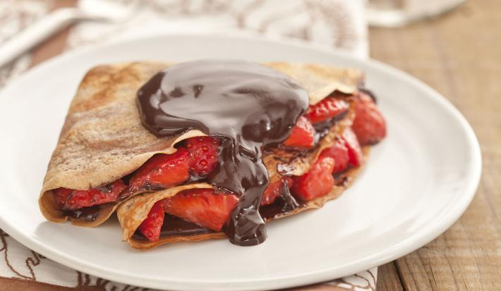 Fotografía en tonos marrones sobre una encimera de madera con un mantel blanco con rayas marrones, un plato blanco poco profundo con la crepe rellena de fresas y chocolate.