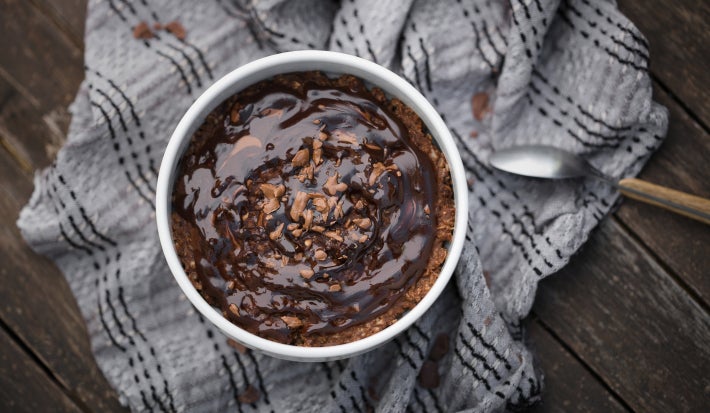 Fotografía de un pastel de avena horneado y frito al aire en un ramequín blanco sobre un paño gris arrugado, colocado sobre una tabla de madera oscura. Se vierte jarabe de chocolate sobre el pastel.