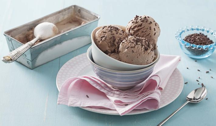 Fotografía en tonos azules y rosas sobre un banco de madera celeste, un paño rosa, un pequeño plato redondo rosa con potes de porcelana encima y bolitas de helado de brigadeiro con chispitas para decorar. Al lado, un botecito con chispitas y una cuchara.