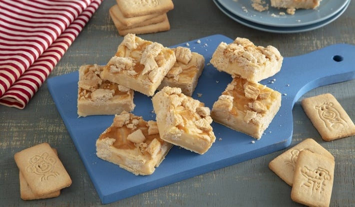 Foto en tonos beige de la receta de dulce para microondas hecho con galletas "Passo", servida en varias porciones sobre una tabla azul con algunas galletas "Passo" de leche alrededor. Al fondo, un mantel a rayas rojas y blancas y un plato azul. Foto en tonos beige de la receta de dulce para microondas hecho con galletas "Passo", servida en varias porciones sobre una tabla azul con algunas galletas "Passo" de leche alrededor. Al fondo, un mantel a rayas rojas y blancas y un plato azul.