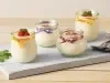 Fotografia em tons de branco e cinza, ao centro  quatro potinhos sobre tábua cinza, decorados com fitas coloridas, frutas, hortelã e raspas de chocolate e contendo mousse, ao lado ramo de hortelã e quatro colheres, tudo sobre bancada de madeira.